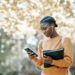 Positive young African American female student with earphones and folder using mobile phone in park