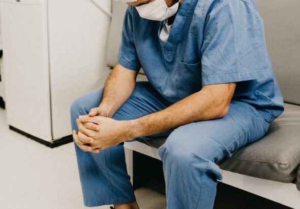 A male doctor in hospital attire sitting pensively, representing healthcare challenges, fraud, doctor, surgeon,