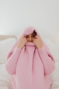 Cozy woman in pink sweater hiding face on a bed, embracing warmth.
